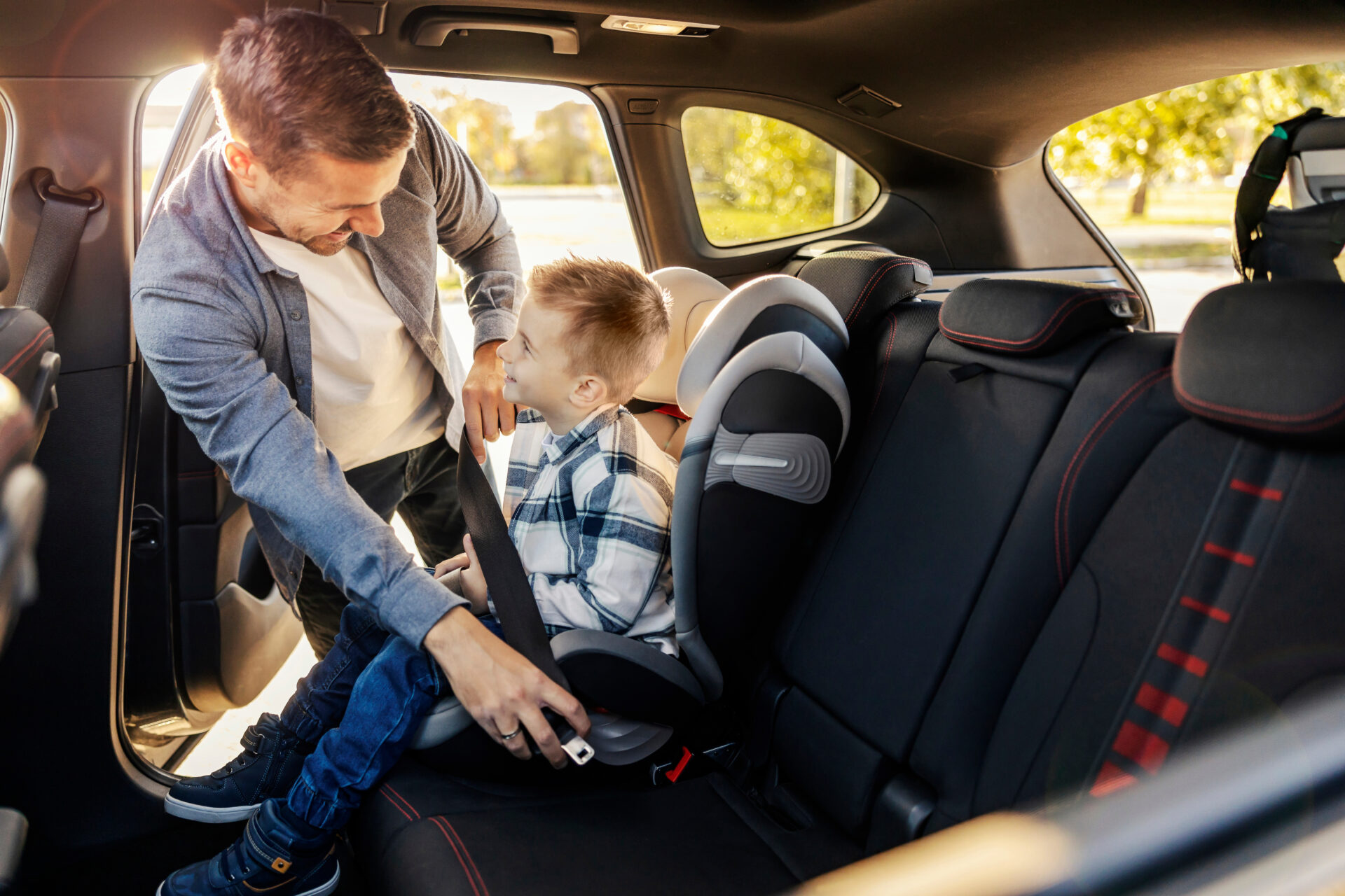 A cheerful dad is buckling up his son in his car seat and preparing for trip.